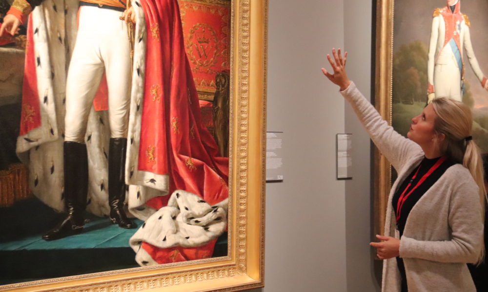 A woman looking at a painting in a museum. She is reaching up to point at something.