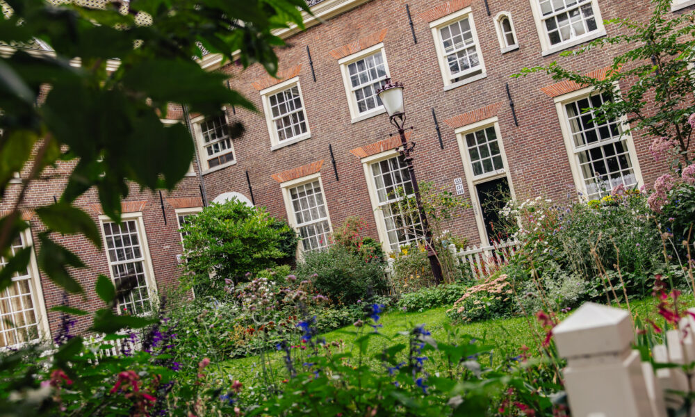 A brick building with white trimmed windows and a garden. The garden is full of flowers and greenery.