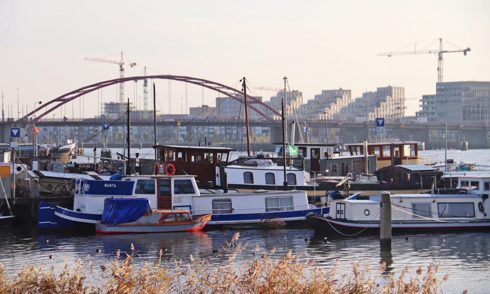 Several boats are moored at a harbour. A bridge and buildings are visible in the background.