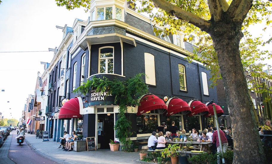 Les clients sont assis devant le café noir Schinkelhafen aux auvents rouges.