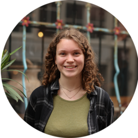 A young woman with curly hair smiling. She is wearing a green shirt and a black plaid shirt.