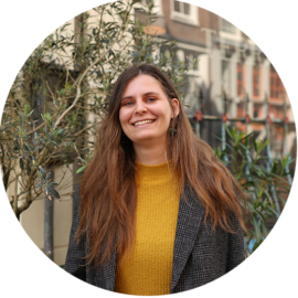 A smiling woman with long brown hair, wearing a yellow sweater and a grey blazer. She appears to be in a city setting.