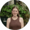 A smiling woman with long brown hair, wearing a brown tank top and a necklace. She is standing in front of green plants.
