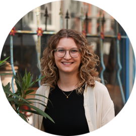 A woman with curly hair and glasses smiling at the camera. She is wearing a black top and a beige cardigan.