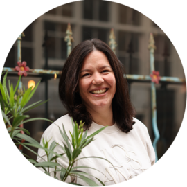 A smiling woman with dark hair standing in front of green plants. She appears to be on a city trip in Amsterdam.