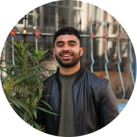 A man with a warm smile wearing a black leather jacket. He is standing in front of a plant and a metal fence.