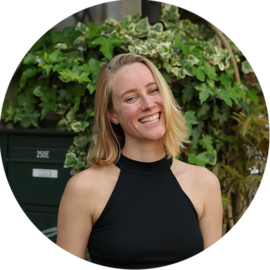 A smiling woman in a black top standing in front of greenery. She appears to be on a city trip in Amsterdam.