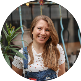 A smiling woman with long hair wearing dungarees. She is outdoors.