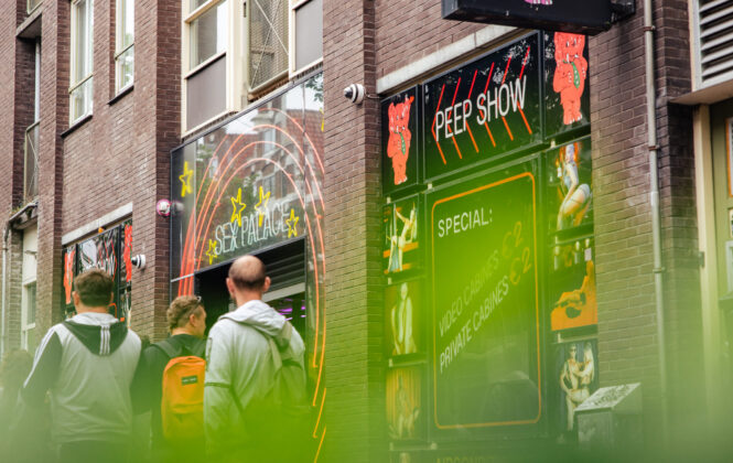 People walking past a colorful entertainment venue in Amsterdam while on a Red Light District tour. A sign reads 'PEEP SHOW'.